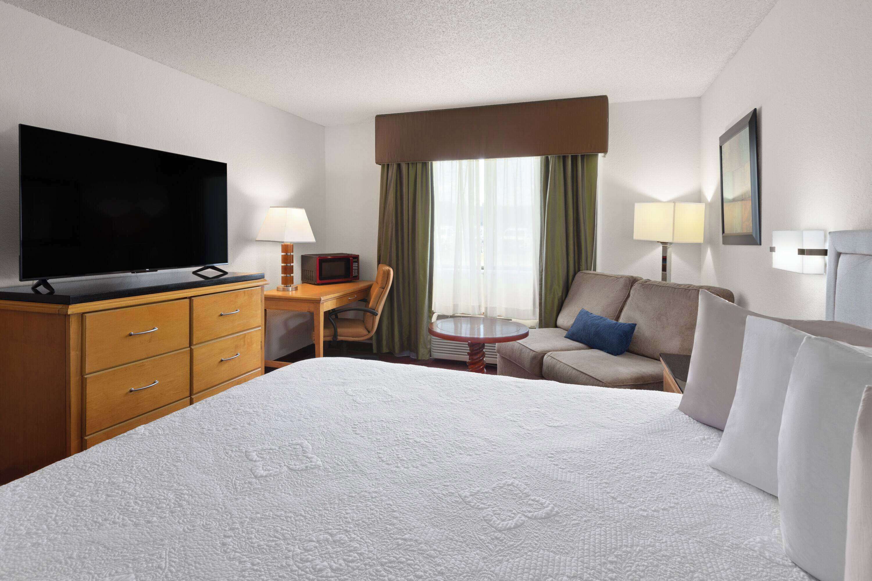 A hotel room with a large bed, a flat-screen TV, a desk with a chair, a small table, and a sofa with pillows.