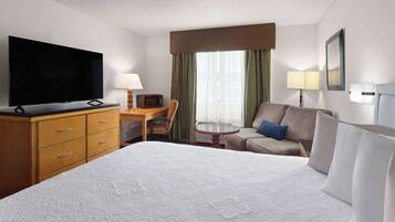 A hotel room with a large bed, a flat-screen TV, a desk with a chair, a small table, and a sofa with pillows.