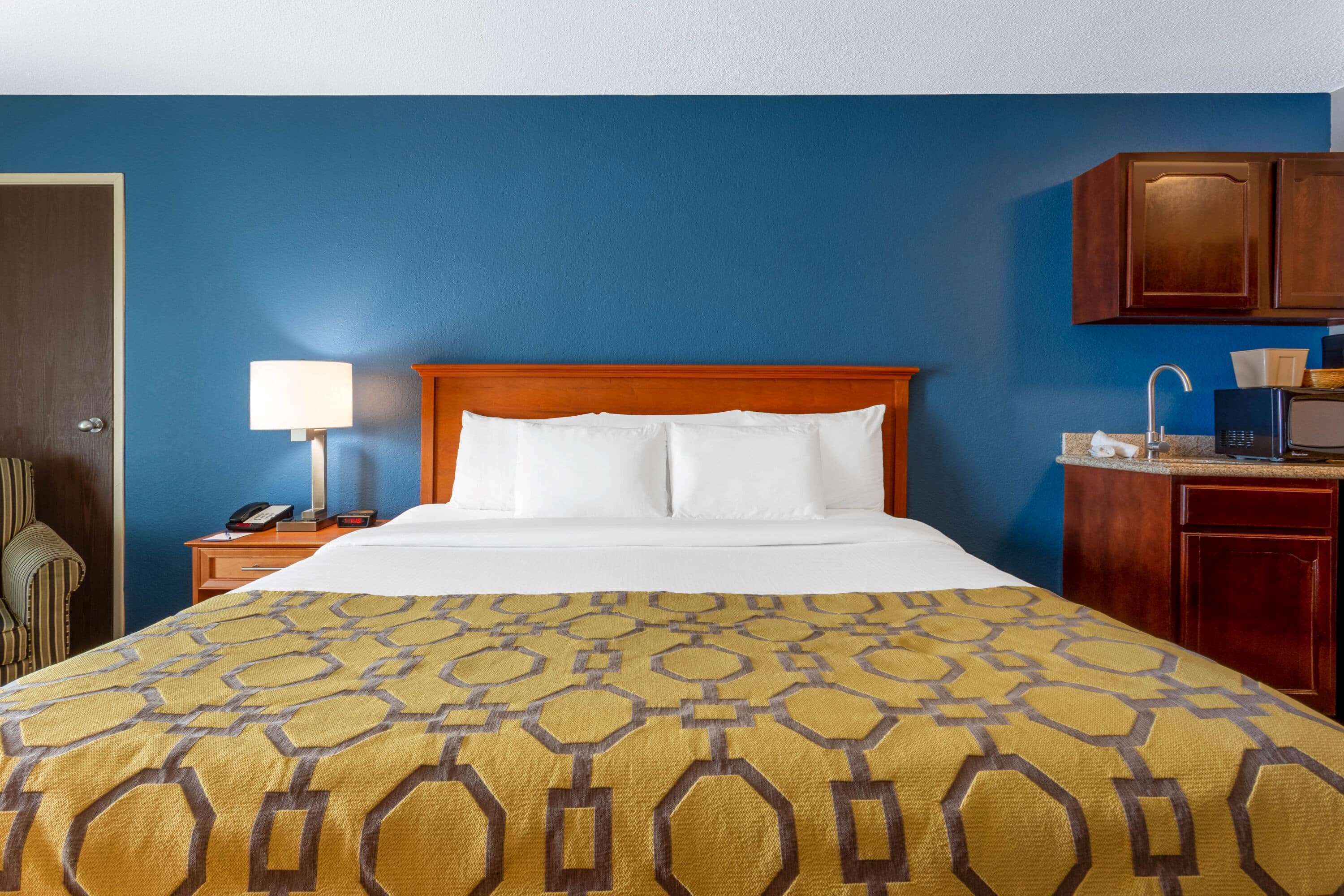 Quarto de hotel com cama, mesa de cabeceira, luminária, cabeceira de madeira, escrivaninha com cadeira e área de cozinha compacta.