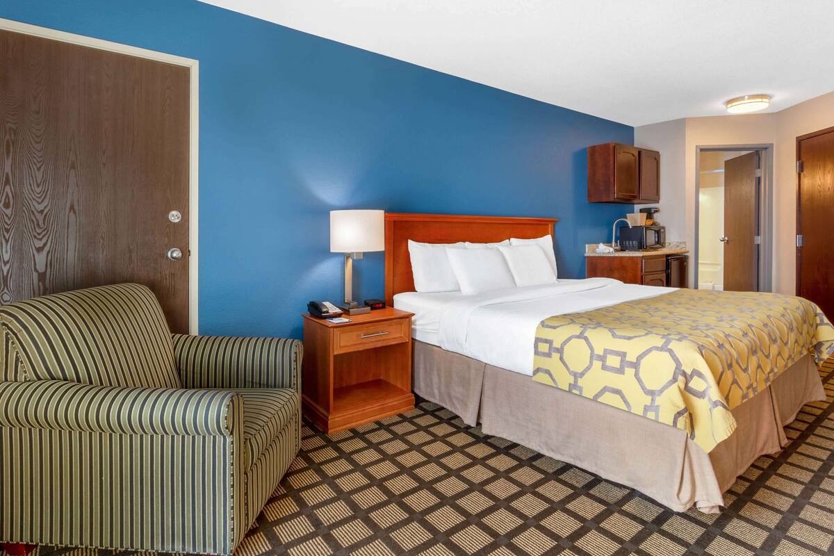 Een hotelkamer met een bed, nachtkastje, stoel, bureau met koffiezetapparaat en een kledingkast.