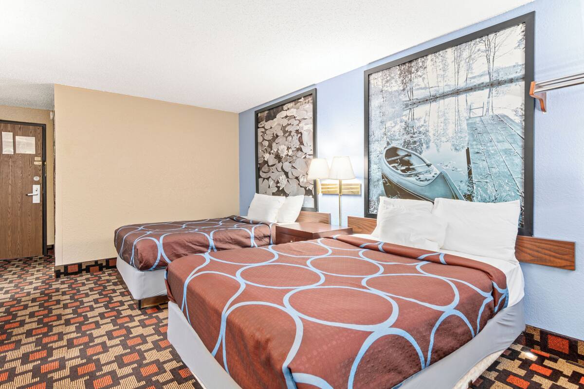 A hotel room with two beds, a wooden headboard, a nightstand with a lamp, and a large window with a winter scene view.