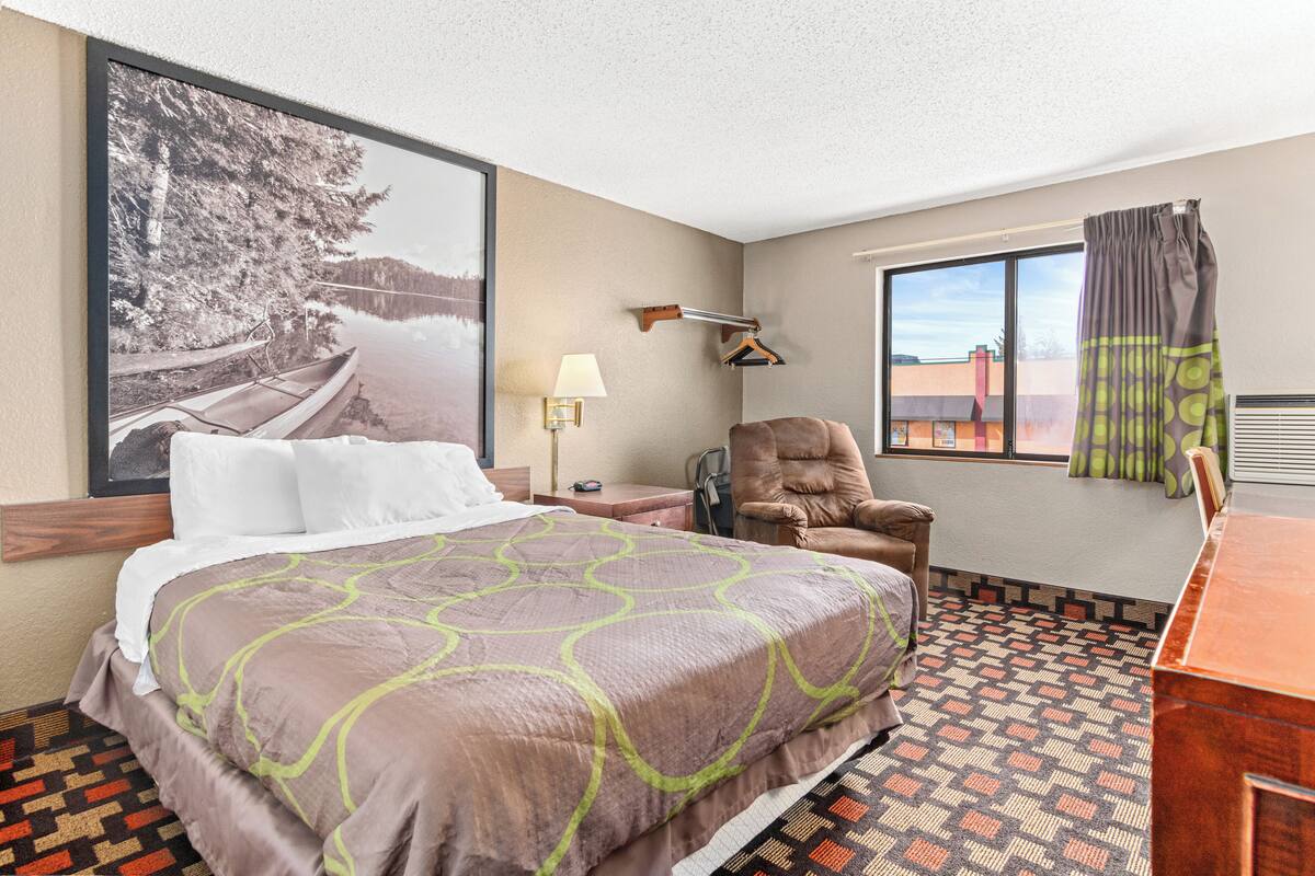 A hotel room with a large bed, a chair, a desk, a window with a view, and a wall-mounted lamp.