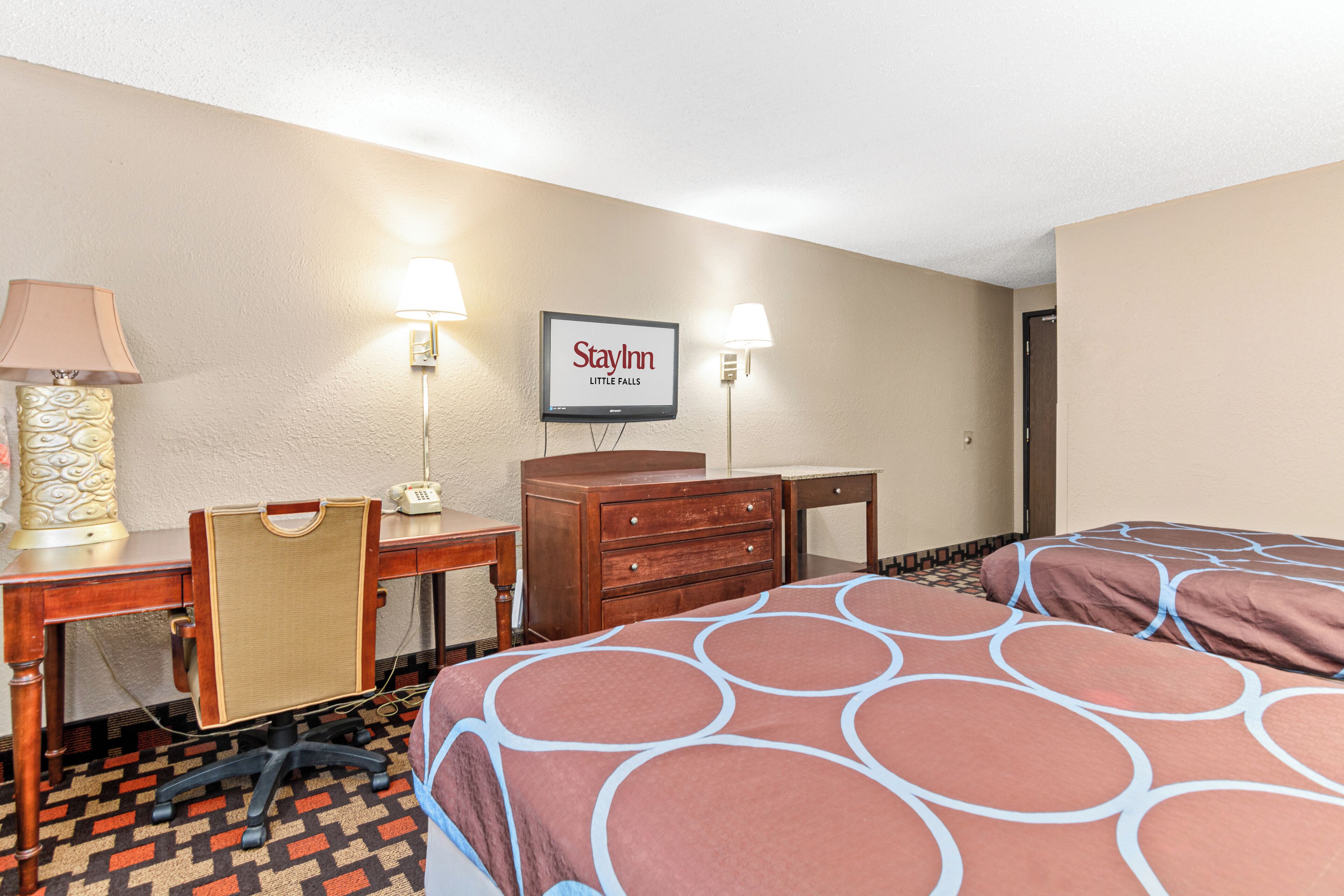 A hotel room with two beds, a desk with a chair, a dresser, and a television mounted on the wall.