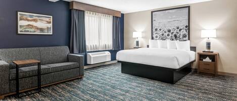 Una habitación de hotel con cama, sofá, lámpara, un cuadro en la pared y ventana con cortinas.
