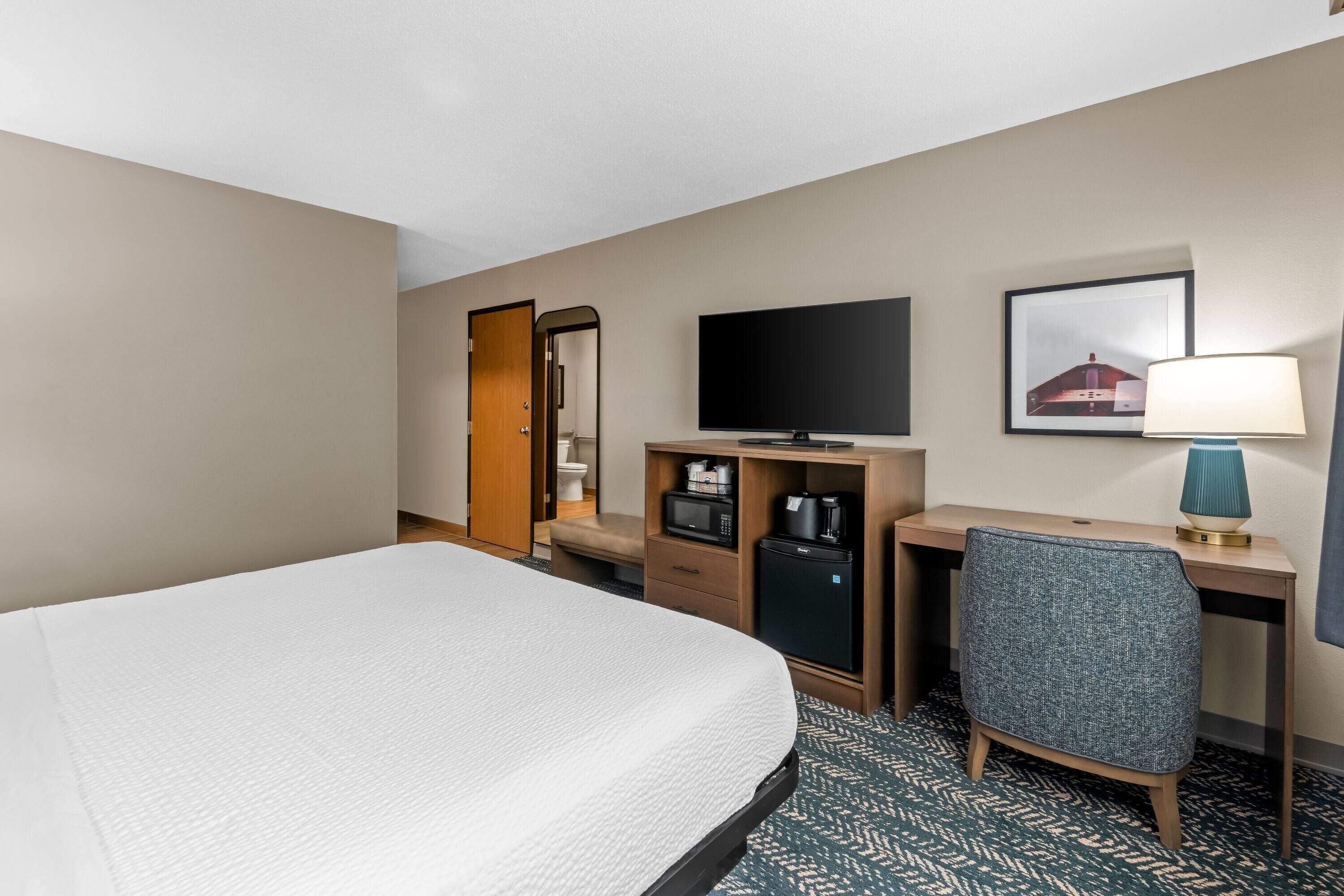 A hotel room with a bed, a desk, a television, a lamp, and a small refrigerator.