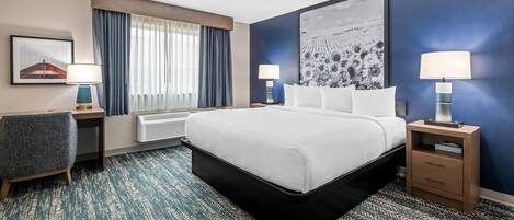 A hotel room with a large bed, a desk, a chair, two bedside tables with lamps, a window with curtains, and a large abstract painting on the wall.