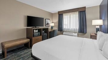 A hotel room with a large bed, a flat-screen TV, a desk with a computer, a bench, and a window with curtains.