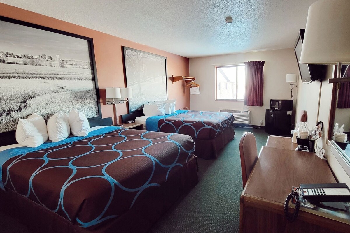 Habitación de hotel con dos camas, un escritorio, una silla, un televisor y un ventanal amplio.