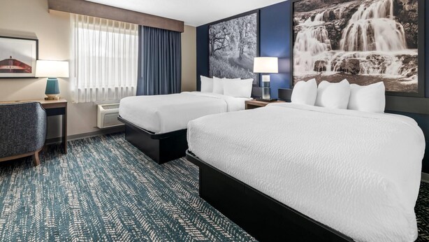 Habitación de hotel con dos camas, un escritorio, una silla, un ventanal con cortinas y un cuadro de una cascada en la pared.
