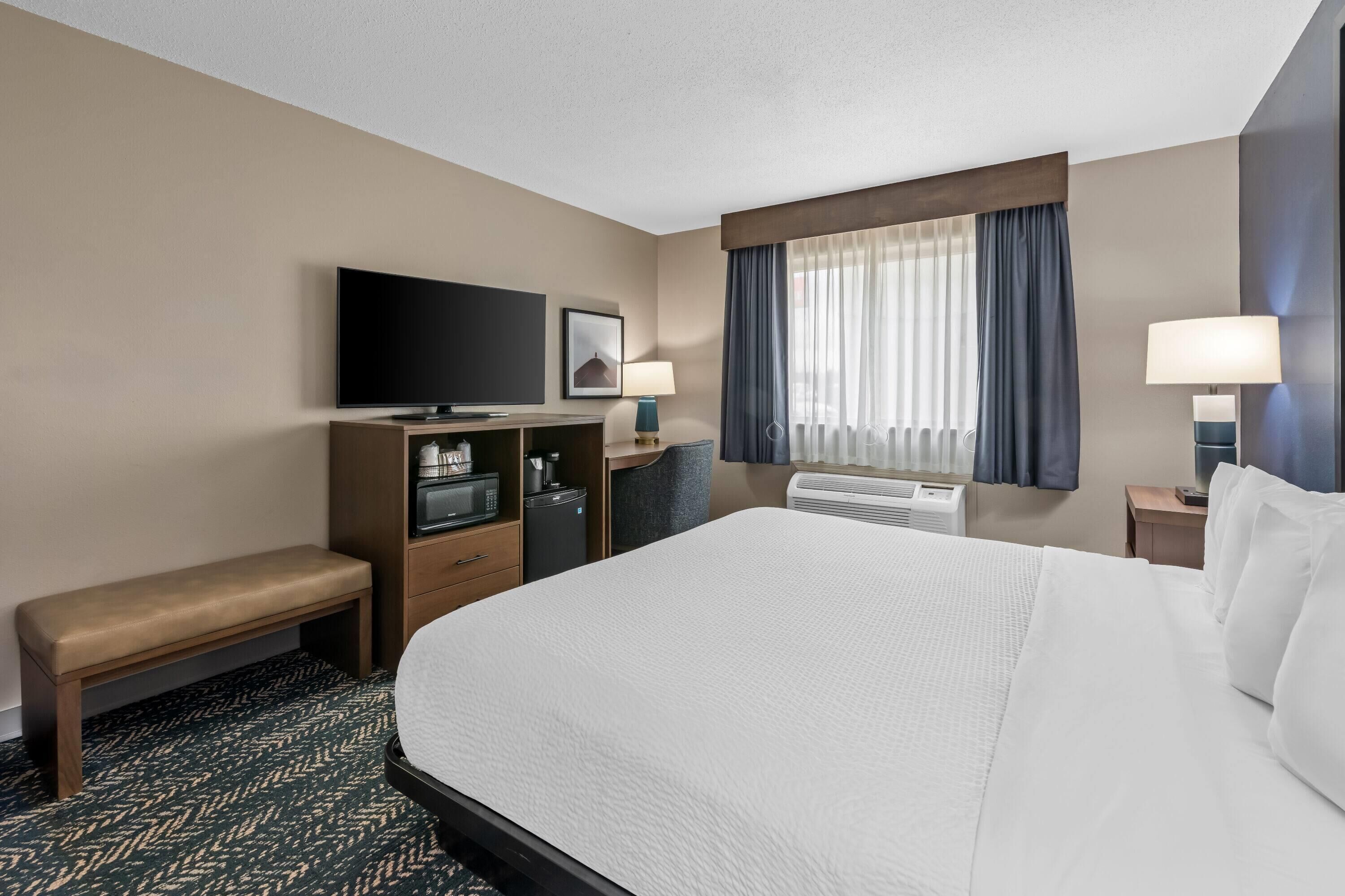 A hotel room with a large bed, a TV, a desk with a computer, a bench, and a window with curtains.