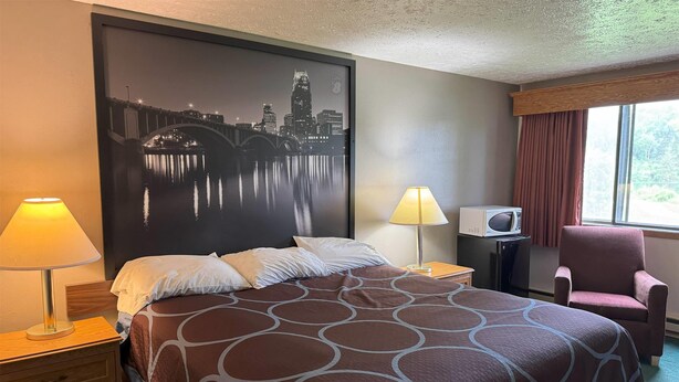 Une chambre d’hôtel avec un lit, une œuvre d’art représentant un paysage urbain accrochée au mur, une lampe, un climatiseur, une chaise et une fenêtre avec des rideaux.