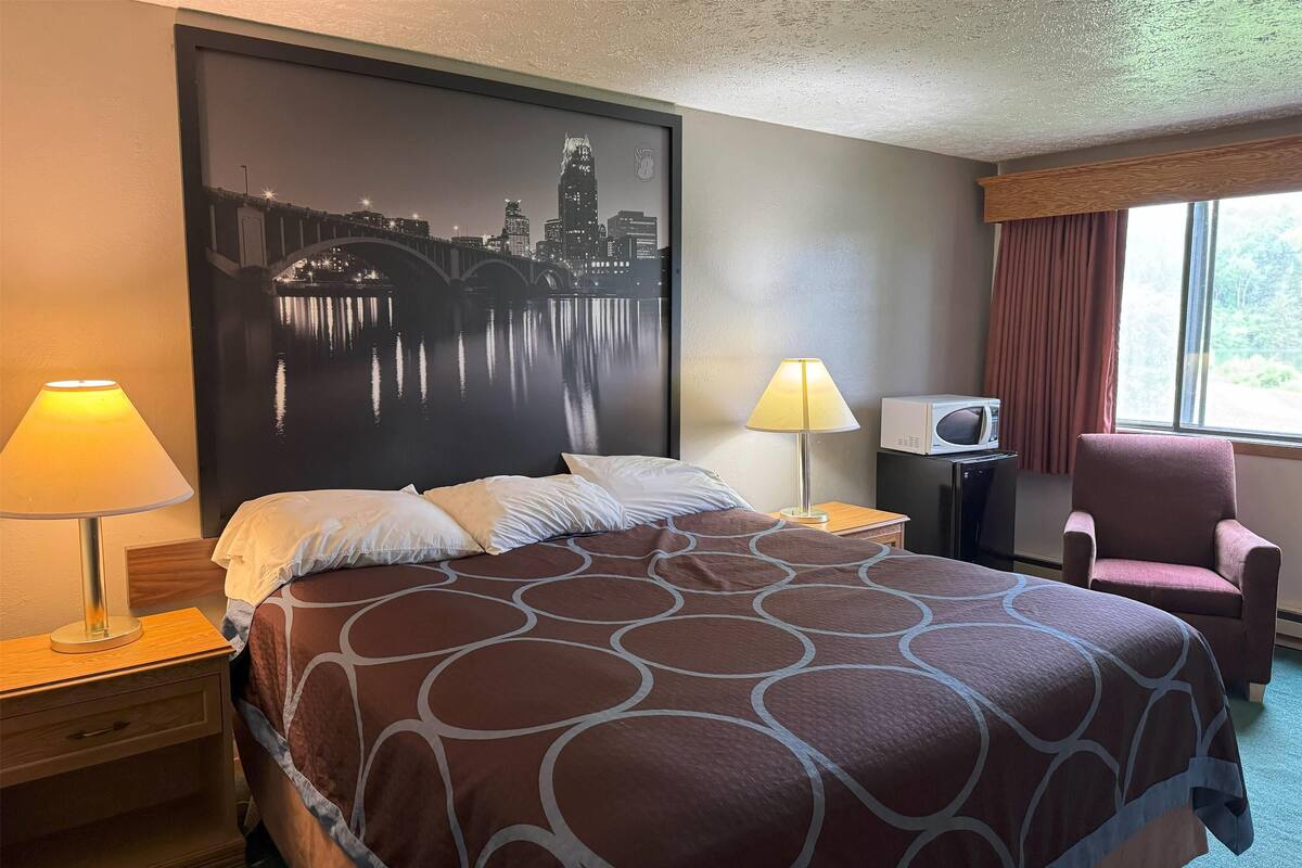 Quarto de hotel com cama, uma pintura de paisagem urbana na parede, luminária, aparelho de ar condicionado, cadeira e janela com cortinas.