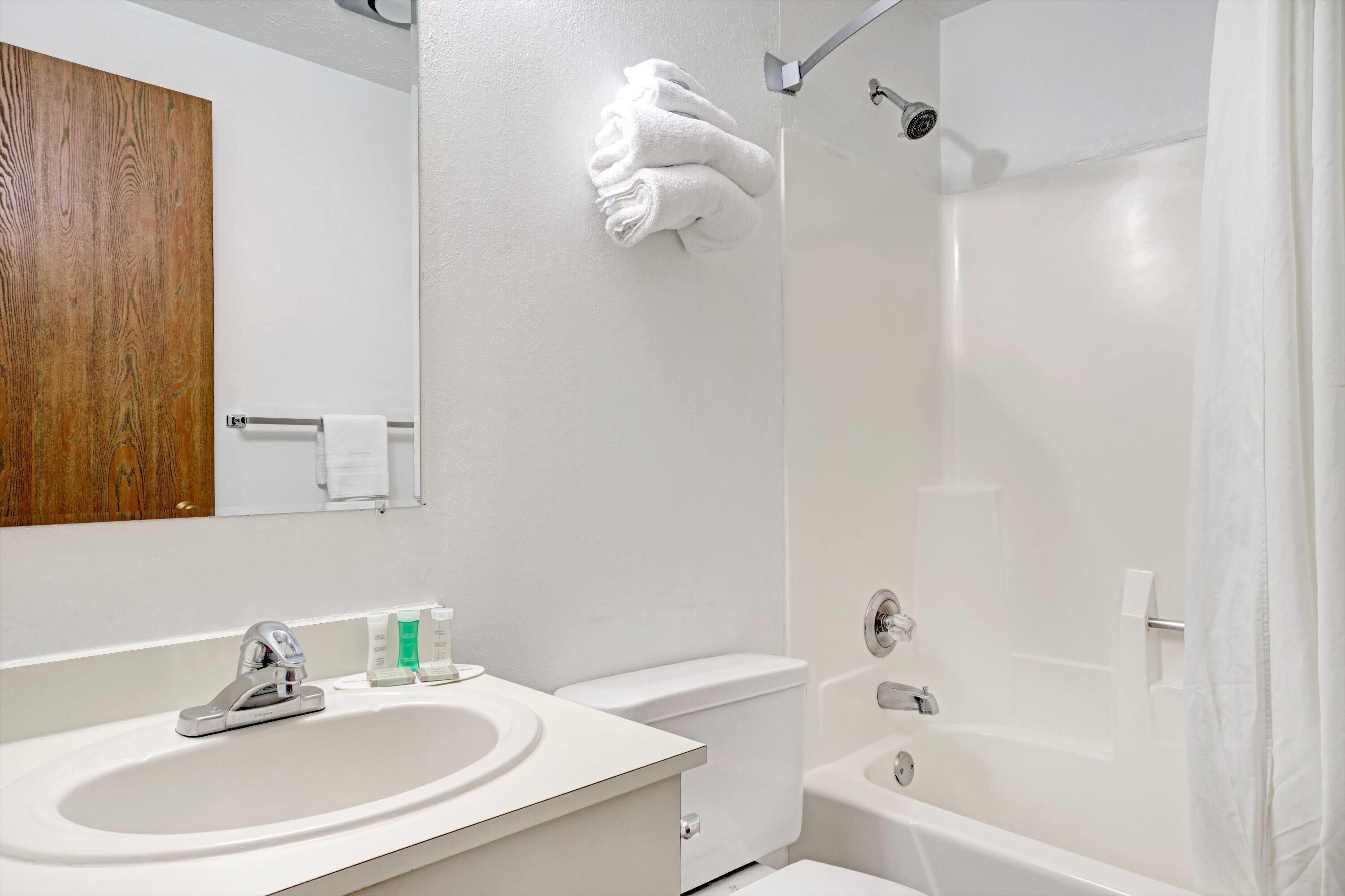 A modern bathroom with a white sink, toilet, and bathtub. There is a towel rack with folded towels and a wooden panel above the mirror.