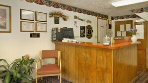 Un comptoir d’accueil avec un ordinateur, un téléphone et un vase de fleurs. Des certificats encadrés sont accrochés au mur, et une pancarte indique : « Accès Internet haut débit gratuit ».