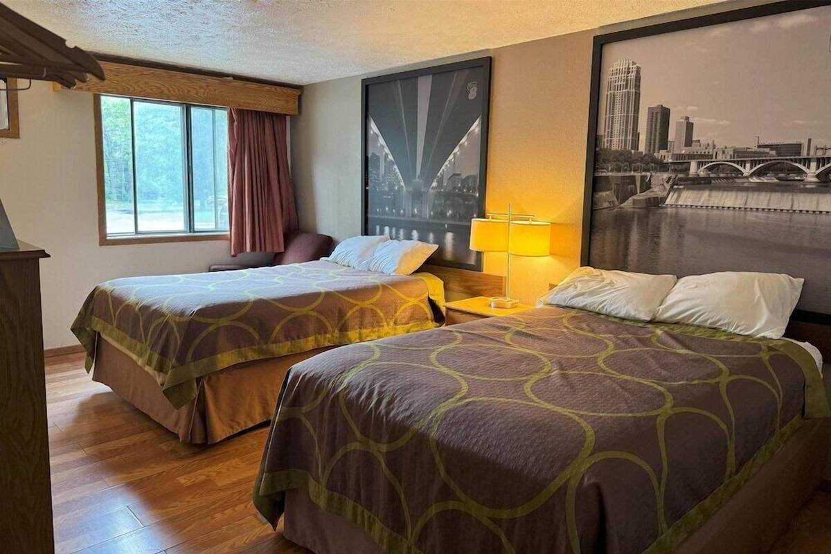 Quarto de hotel com duas camas, janela com cortinas, luminária e pintura de paisagem urbana na parede.