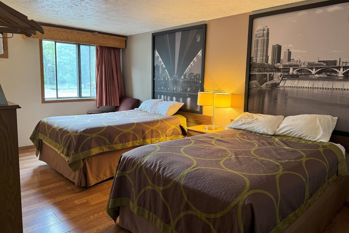 Quarto de hotel com duas camas, janela com cortinas, luminária e um pôster de paisagem urbana na parede.