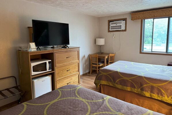Habitación de hotel con dos camas, un escritorio con silla, televisor, microondas, refrigerador y ventana con unidad de aire acondicionado.