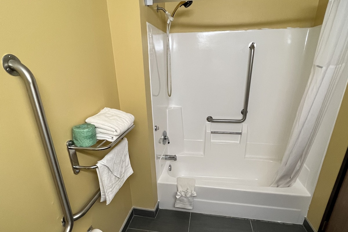 Un baño con bañera, ducha e inodoro. Cuenta con barras de apoyo para mayor seguridad.