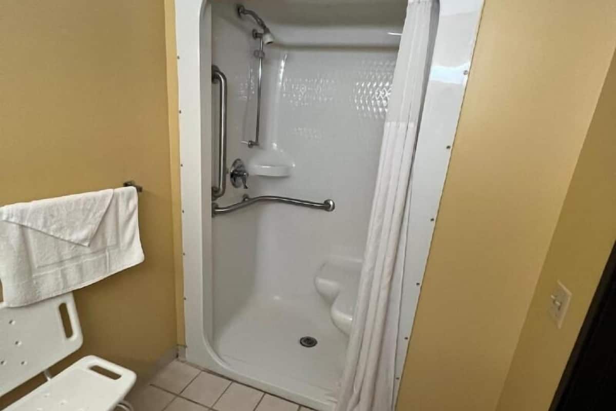 Un baño pequeño con ducha, una barra de apoyo, un banco blanco y un perchero para toallas.