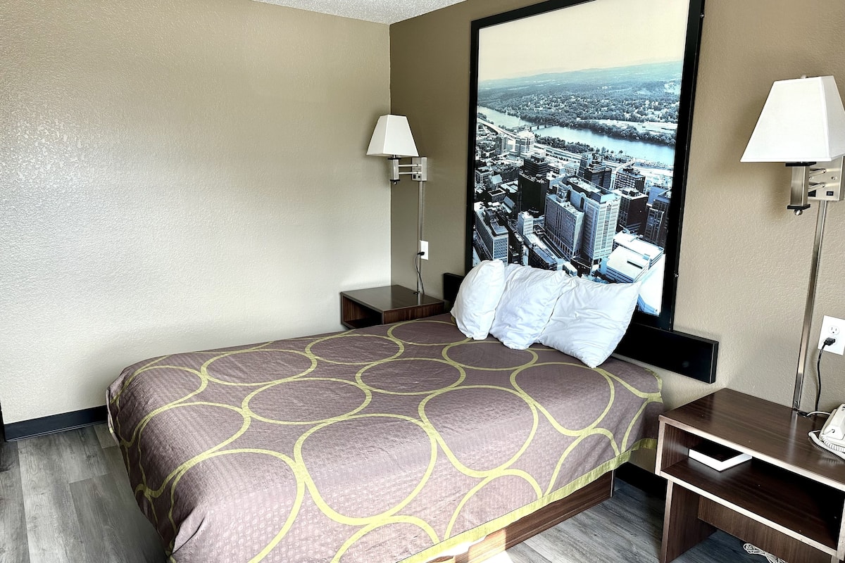 Une chambre d’hôtel avec un grand lit, deux lampes de chevet, une table de chevet et un téléviseur fixé au mur qui diffuse des images d’un paysage urbain.
