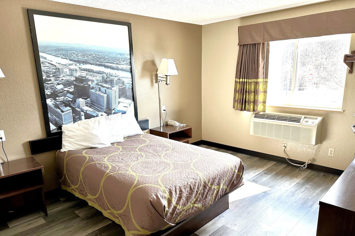 Une chambre d’hôtel comprenant un lit, une table de chevet, une lampe, une fenêtre avec des rideaux et un appareil de climatisation.