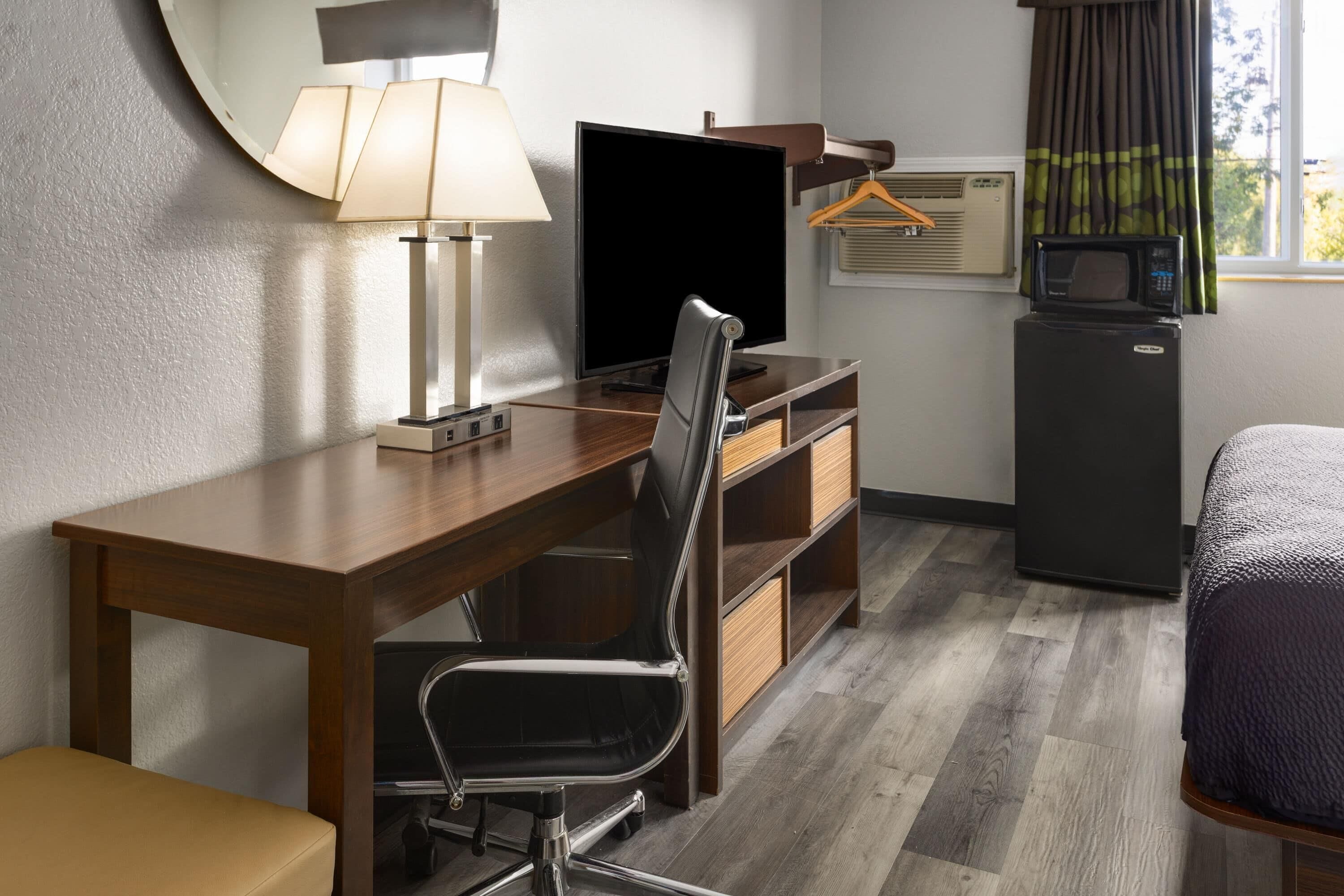 A hotel room with a wooden desk, a chair, a television, a small refrigerator, a microwave, a hanging clothes rack, and a bed.
