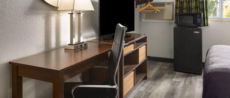 A hotel room with a wooden desk, a chair, a television, a small refrigerator, a microwave, a hanging clothes rack, and a bed.