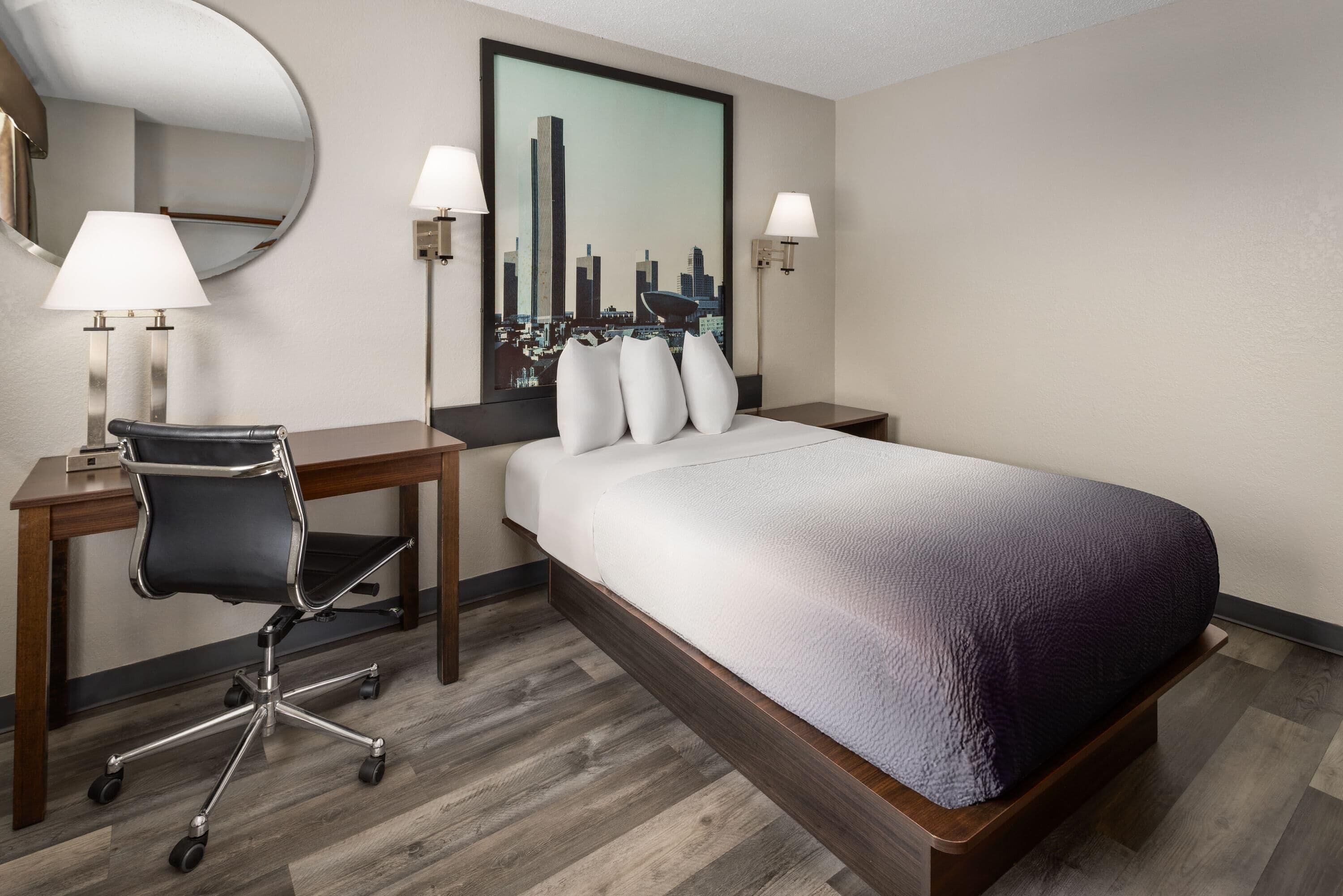 Habitación de hotel con cama, escritorio, silla, lámparas y un cuadro de paisaje urbano.