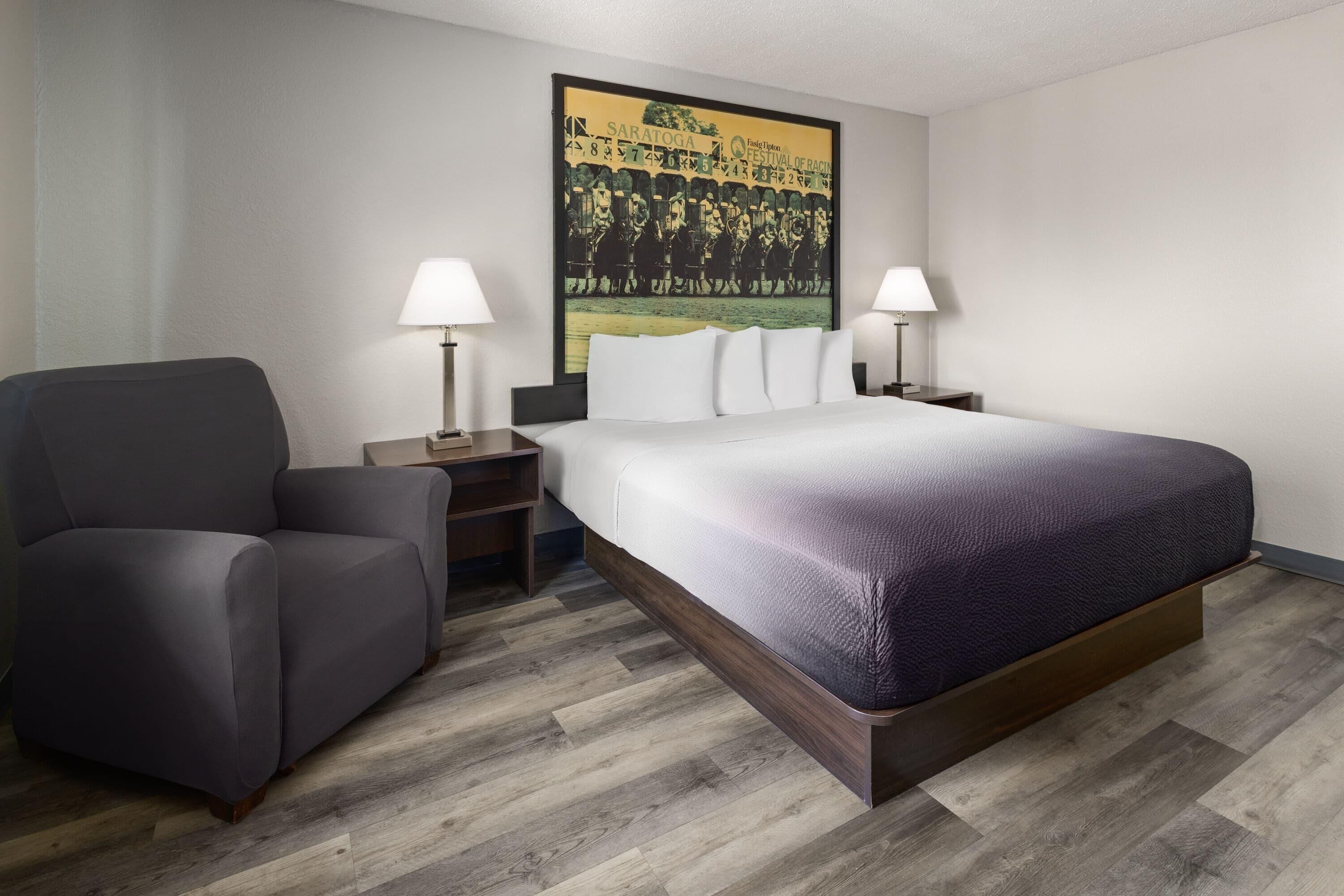 A hotel room with a large bed, two bedside tables with lamps, a gray armchair, and a wall-mounted artwork.