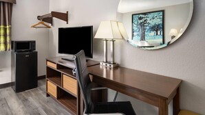 Une chambre d’hôtel équipée d’un bureau en bois, d’un téléviseur à écran plat, d’un mini-réfrigérateur, d’une chaise, d’une lampe et d’un miroir rond fixé au mur.