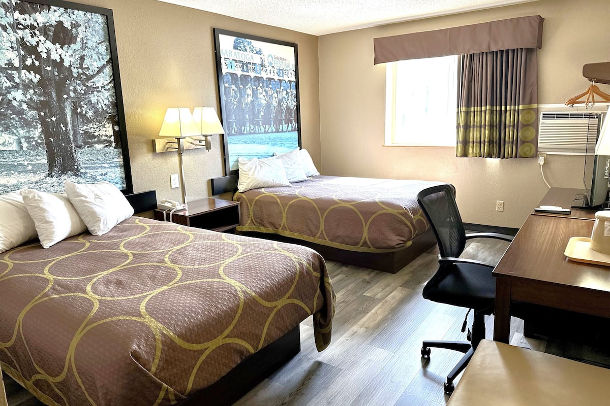 Une chambre d’hôtel avec deux lits, un bureau, une chaise, une lampe, une affiche et une fenêtre avec des rideaux.
