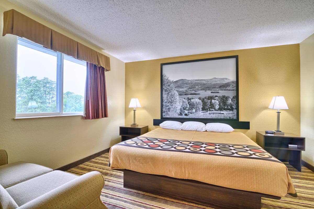 Une chambre d’hôtel avec un grand lit, deux tables de chevet, une chaise, une fenêtre avec des rideaux et un tableau représentant un paysage accroché au mur.