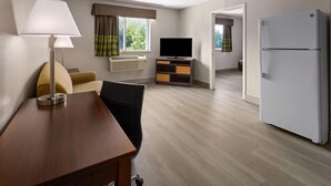 Une chambre d’hôtel avec une table à manger en bois, un canapé, une télévision, un réfrigérateur et une fenêtre avec des rideaux.