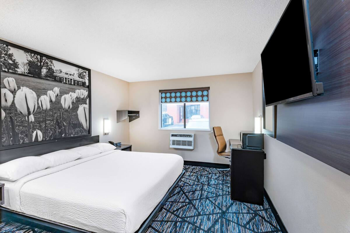 A hotel room with a large bed, a flat-screen TV, a window with blinds, a desk with a chair, and a wall-mounted air conditioning unit.