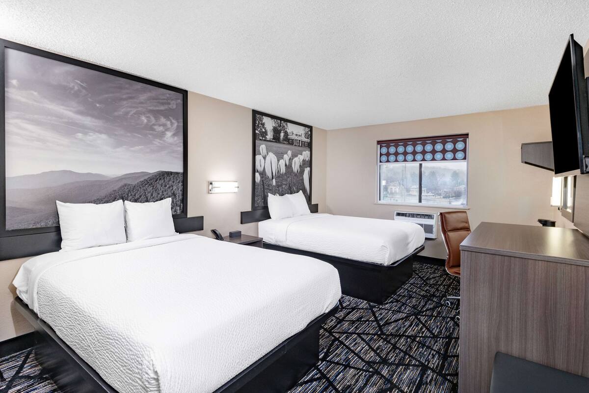 A hotel room with two beds, a desk, a chair, a TV, and a window with blinds.