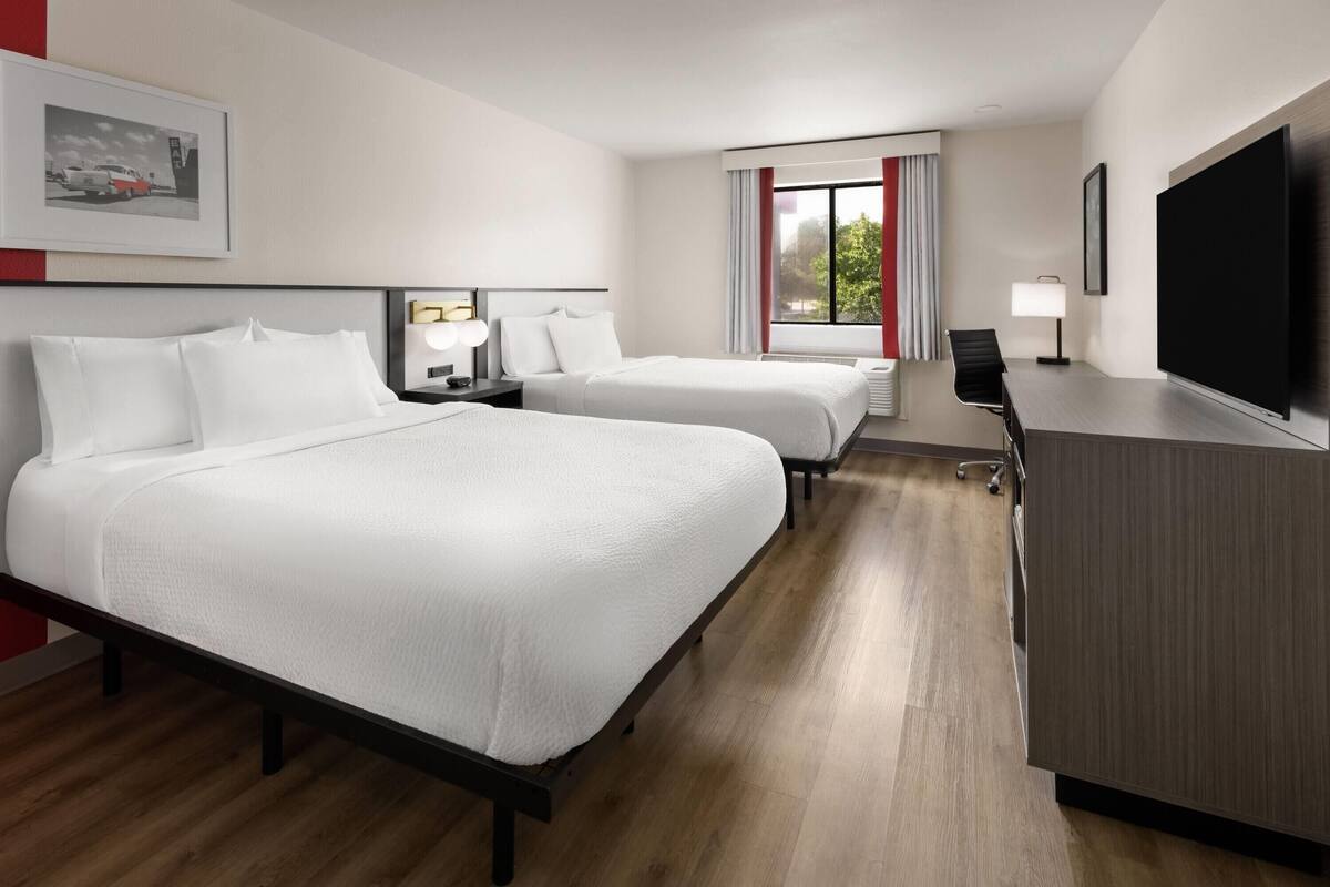 A hotel room with two beds, a desk, a TV, and a window with curtains.