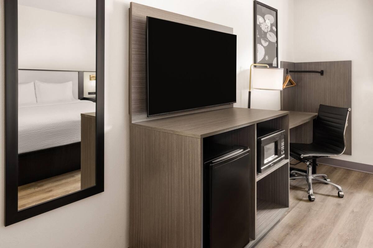 A modern hotel room with a bed, a flat-screen TV, a desk with a computer, a microwave, and a chair.