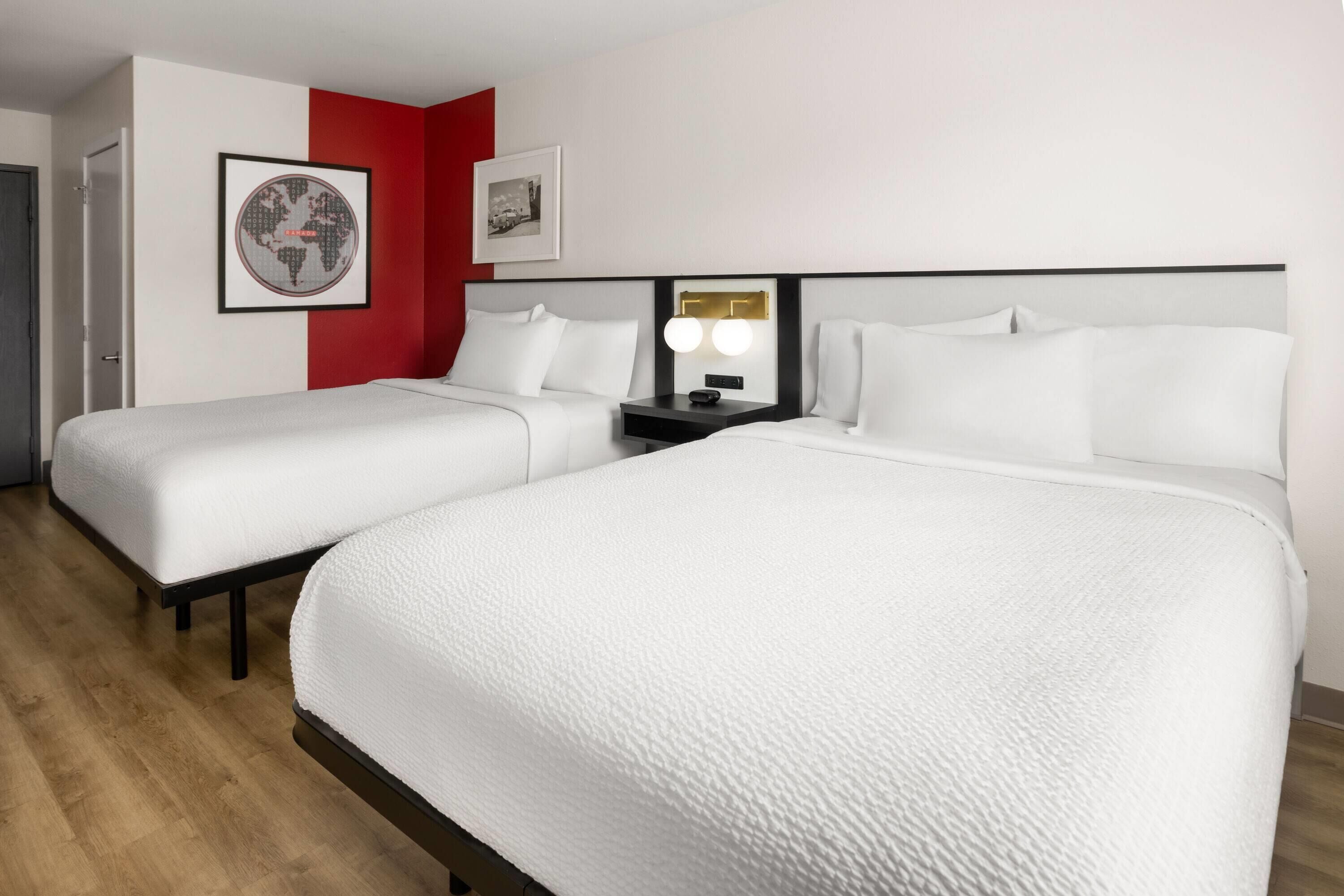 A hotel room with two beds, a red accent wall, a framed world map, and a nightstand with a lamp.
