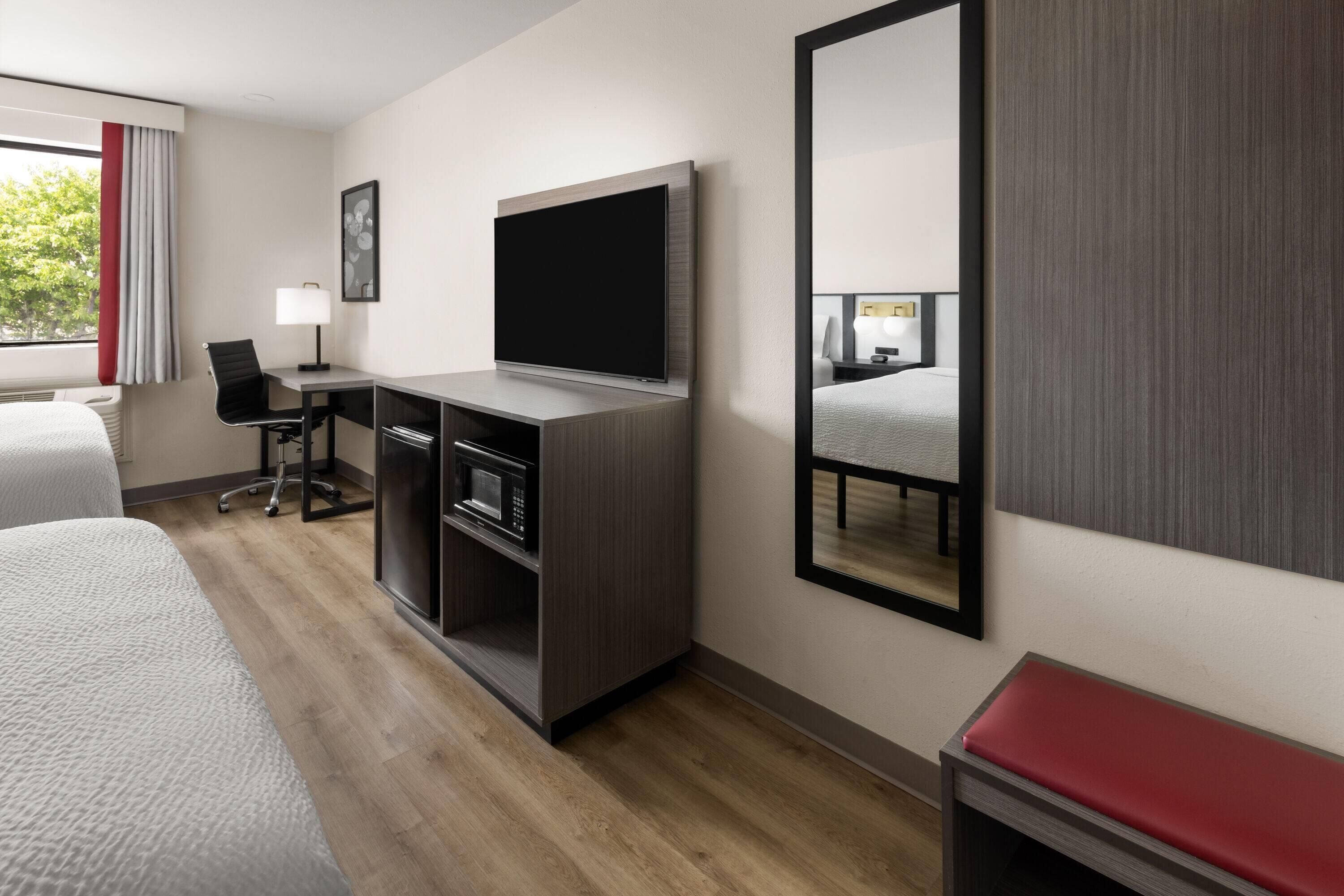 A hotel room with a bed, a desk with a TV, a mirror, and a red bench.