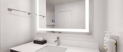 Un baño moderno con un lavabo blanco, un espejo grande con iluminación integrada, un perchero y un dispensador de jabón.