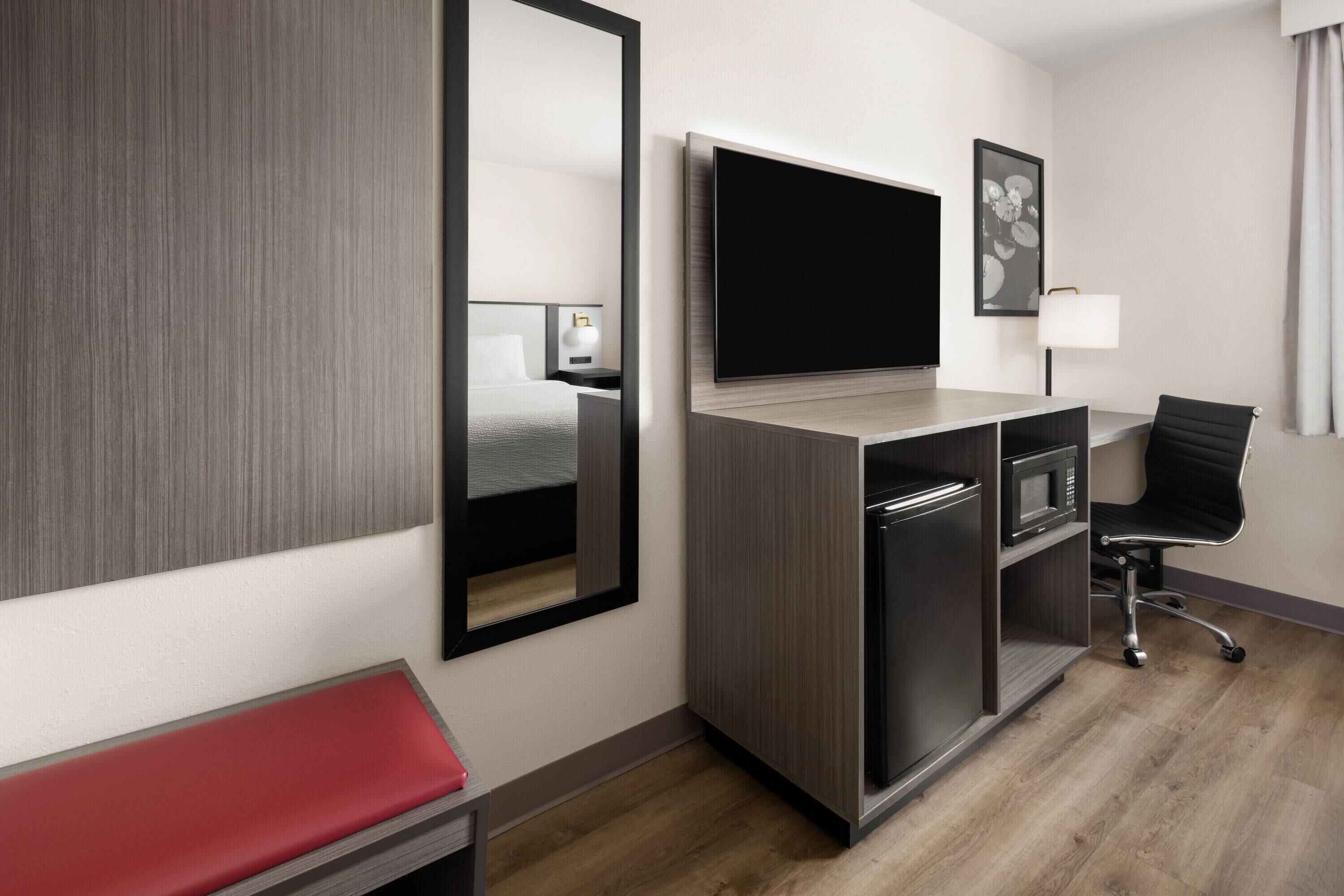 Ein modernes Hotelzimmer mit einem großen Flachbildfernseher, einem Schreibtisch mit Stuhl, einer Mikrowelle und einer roten Bank.