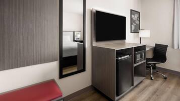 A modern hotel room with a large flat-screen TV, a desk with a chair, a microwave, and a red bench.