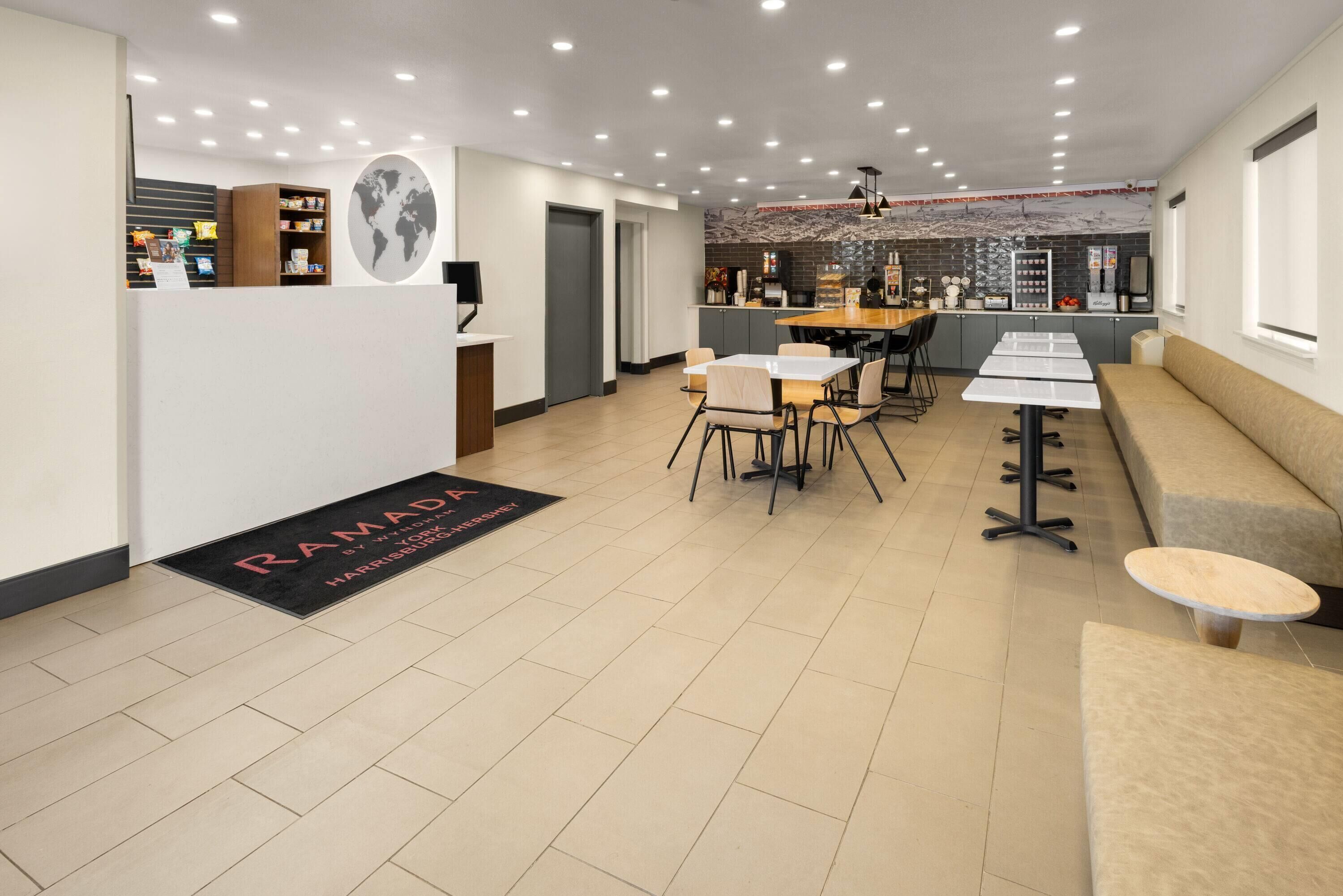 Un vestíbulo moderno de hotel con recepción, un área de comedor con mesas y sillas, una cocina y un espacio de descanso con un sofá y una pequeña mesa redonda.