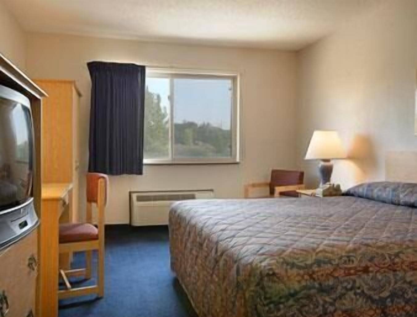 A hotel room with a bed, a desk with a chair, a window with curtains, and a heater.