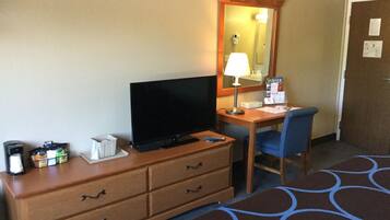 A hotel room with a wooden desk, a television, a chair, a mirror, a coffee maker, and a bed with a patterned bedspread.