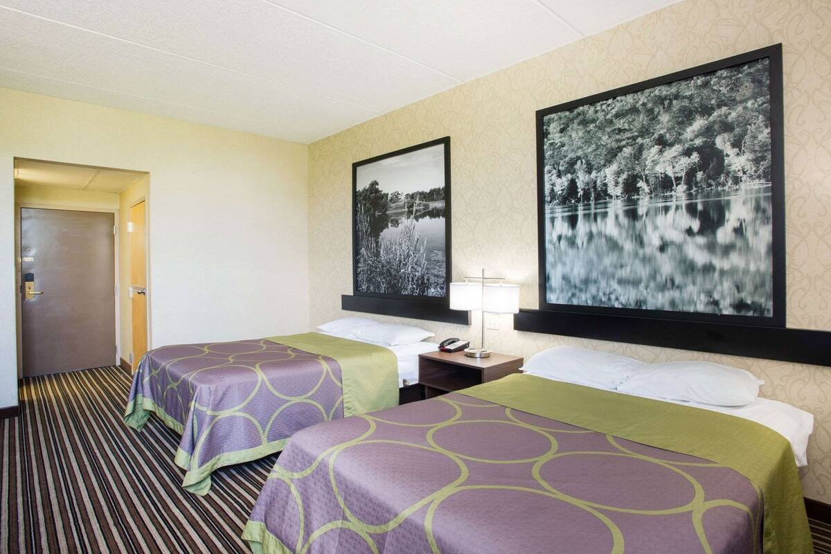 A hotel room with two beds, a nightstand, a lamp, and two large landscape paintings on the walls.
