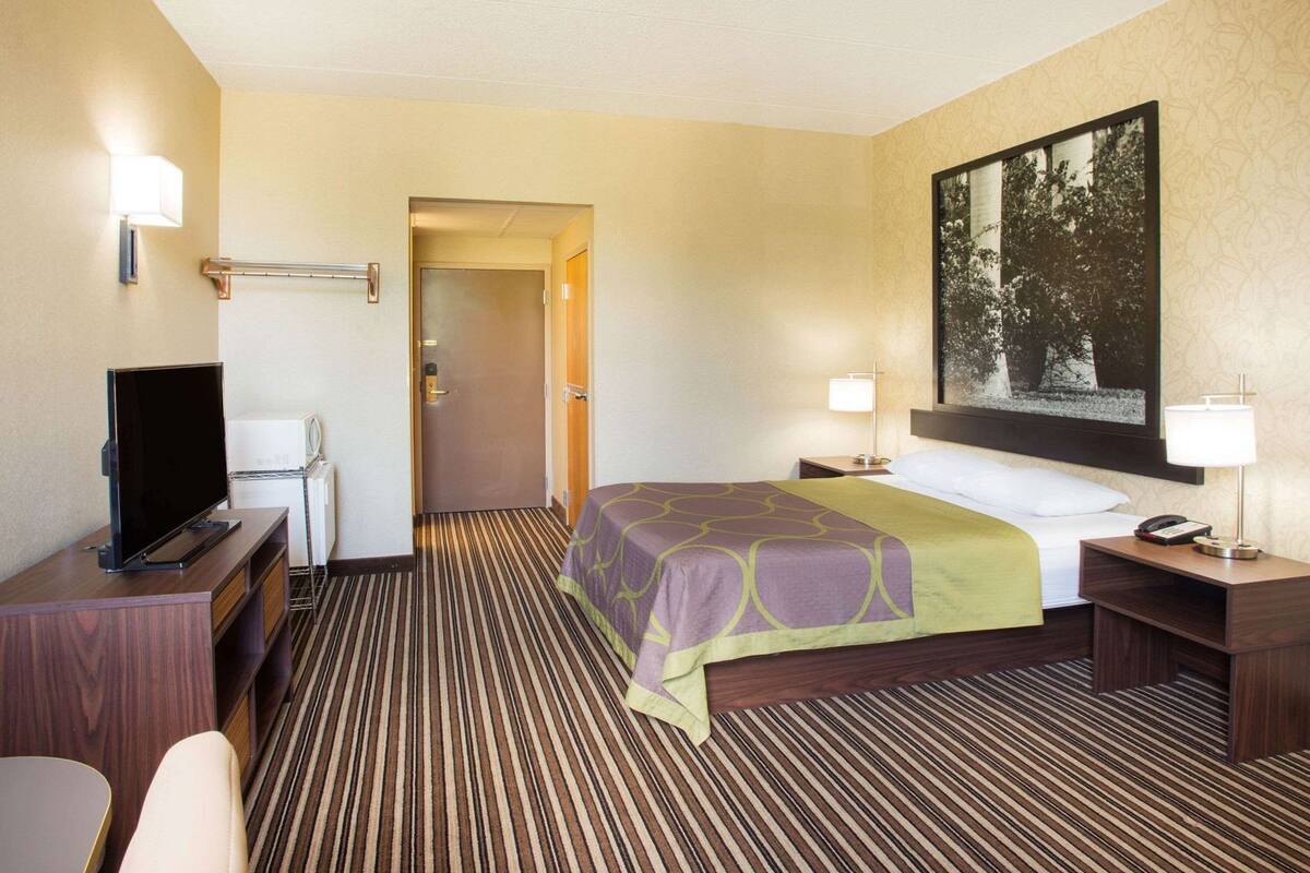 A hotel room with a bed, a desk with a television, a bedside table with a lamp, and a framed picture on the wall.