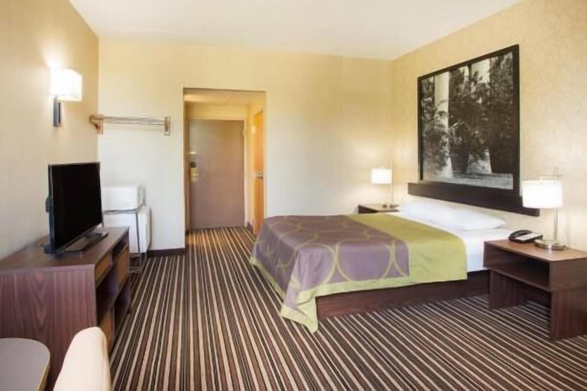 A hotel room with a bed, a desk with a television, a chair, a desk lamp, and a painting on the wall.