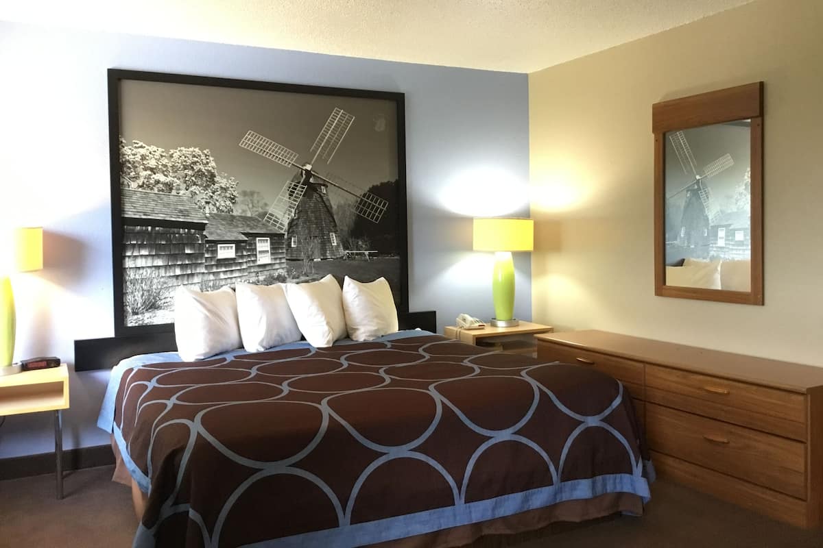 Ein Hotelzimmer mit einem großen Bett, einem Nachttisch mit Lampe, einer hölzernen Kommode und einem an der Wand befestigten Spiegel.