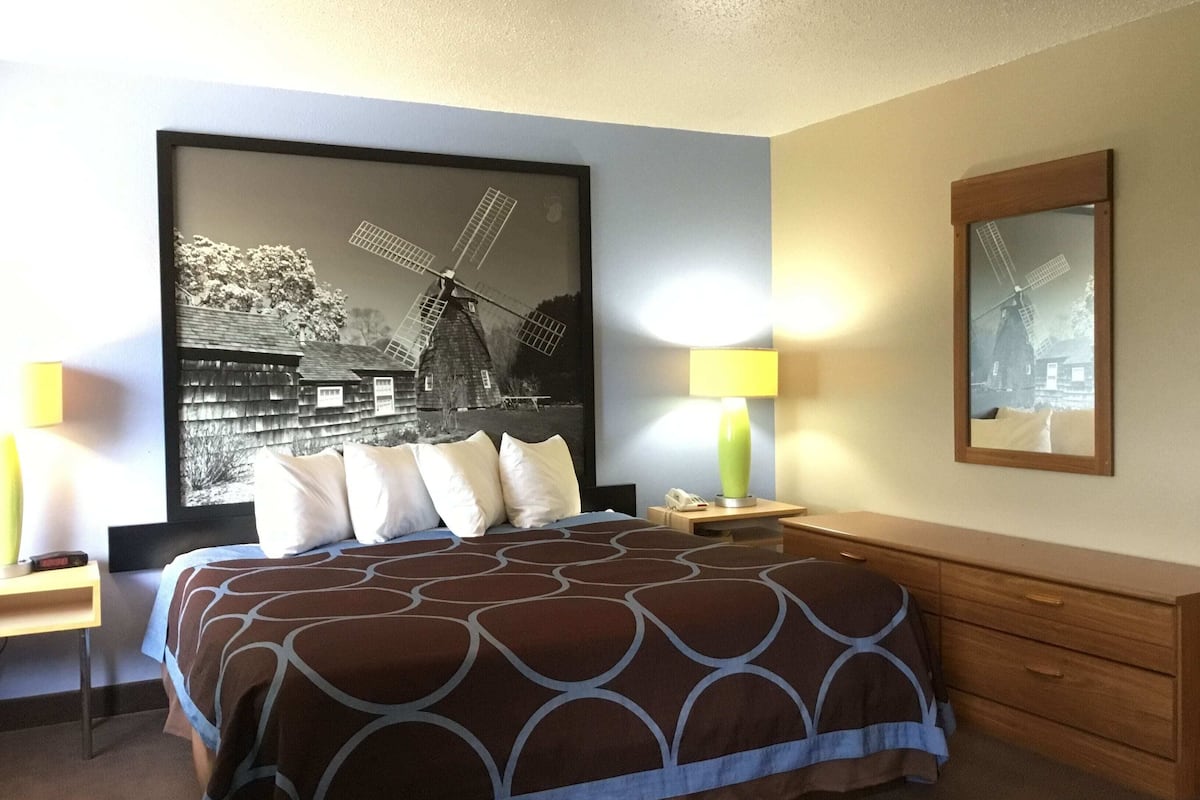 Ein Hotelzimmer mit Bett, Nachttischlampen, einem Nachttisch, einer Holzkommode und einem großen gerahmten Bild einer Windmühle.